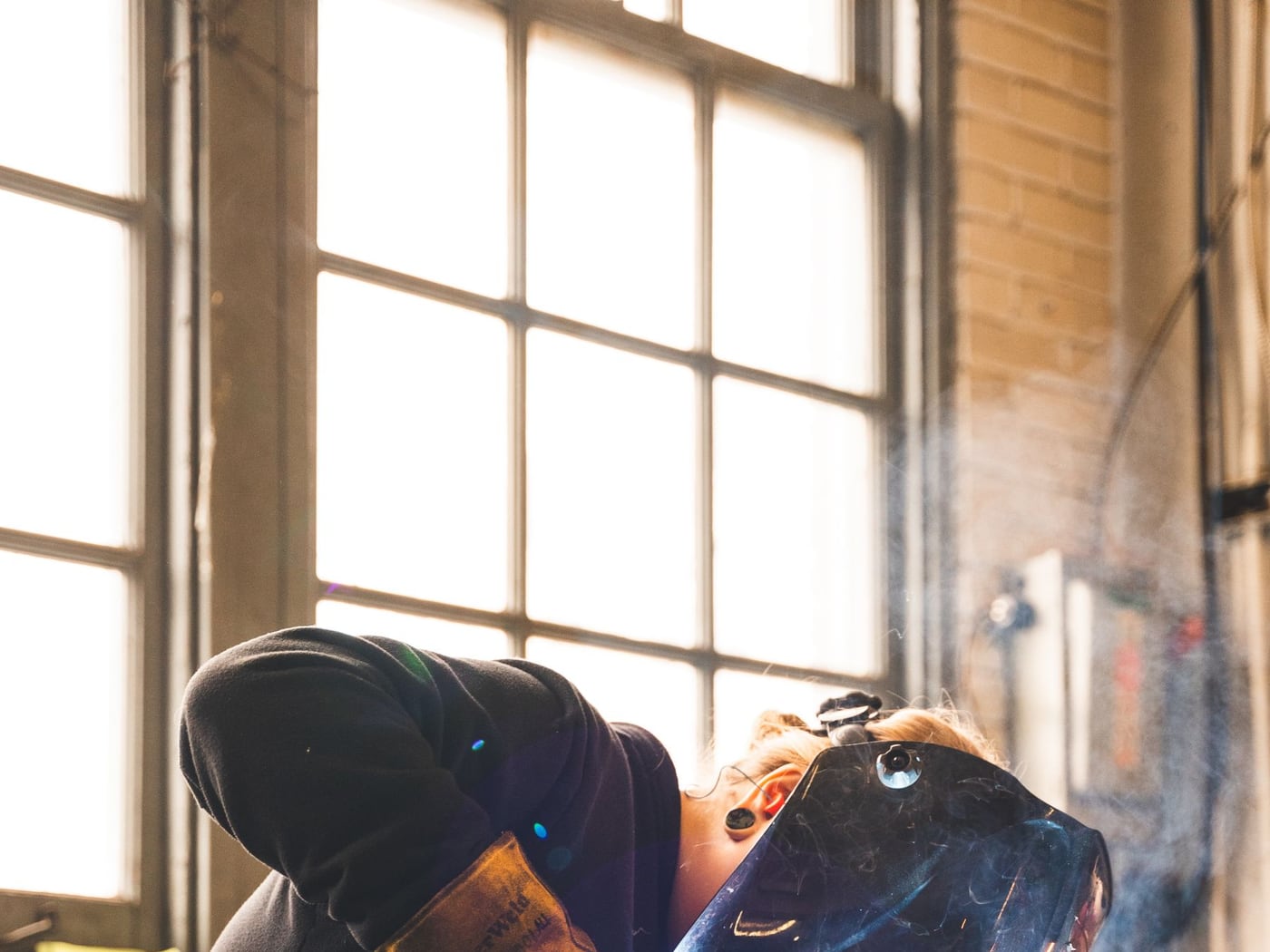 A welder in a helmet running a MIG gun along steel on a bench, lit by a tall shop window &mdash; representative imagery, not a specific Late Night Fabrication job