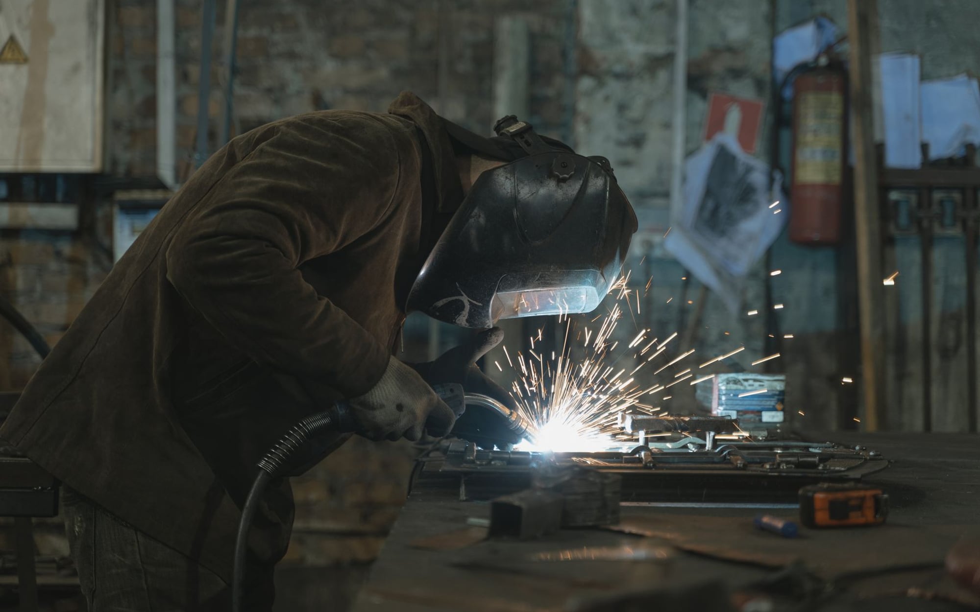 A welder in a full welding helmet working over steel in a dark shop, sparks scattering — representative imagery, not a specific Late Night Fabrication job